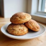 Three soft, puffy cinnamon cookies generously coated in cinnamon sugar, stacked on a white plate.