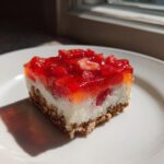 A square slice of strawberry pretzel salad with a pretzel crust, cream cheese layer, and bright red strawberry topping.