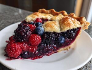 A close-up of a generous slice of triple berry pie showing juicy raspberries, blackberries, and blueberries.