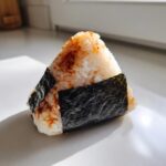 A triangular tuna onigiri rice ball wrapped partially in dark nori seaweed and sprinkled with seasoning.