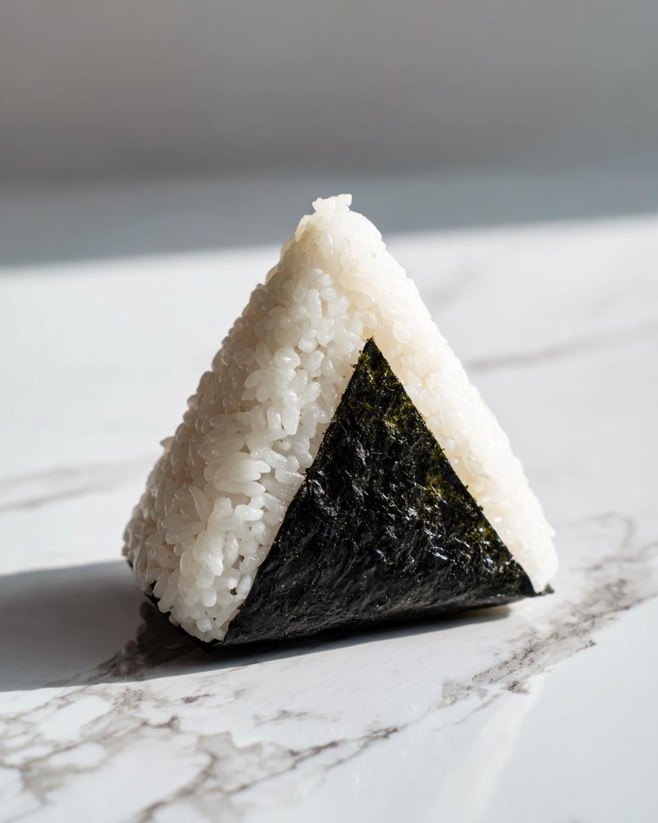 A single, triangular tuna onigiri wrapped partially in dark nori seaweed, resting on a white marble surface.