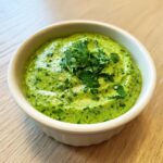 Close-up of a vibrant green cilantro lime sauce garnished with fresh cilantro leaves in a small white bowl.