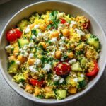 Close-up of a colorful quinoa salad featuring yellow quinoa, chickpeas, cucumber chunks, cherry tomatoes, and crumbled feta cheese.