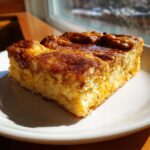 A square slice of golden brown baked french toast topped with cinnamon sugar, served on a white plate.