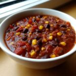 Close-up of a white bowl filled with rich, thick black bean chili featuring visible chunks of tomato and bright yellow corn kernels.