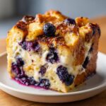 A close-up of a square slice of rich Blueberry French Toast Casserole showing toasted bread cubes, creamy filling, and bursting blueberries.