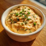 A close-up of creamy, orange-hued remoulade sauce topped with fresh chopped green onions in a white ramekin.