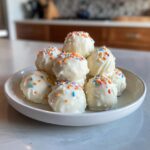 A pile of white chocolate coated sugar cookie truffles topped with colorful sprinkles on a white plate.
