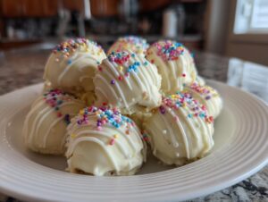 A plate piled high with white chocolate coated sugar cookie truffles, drizzled with white icing and colorful sprinkles.